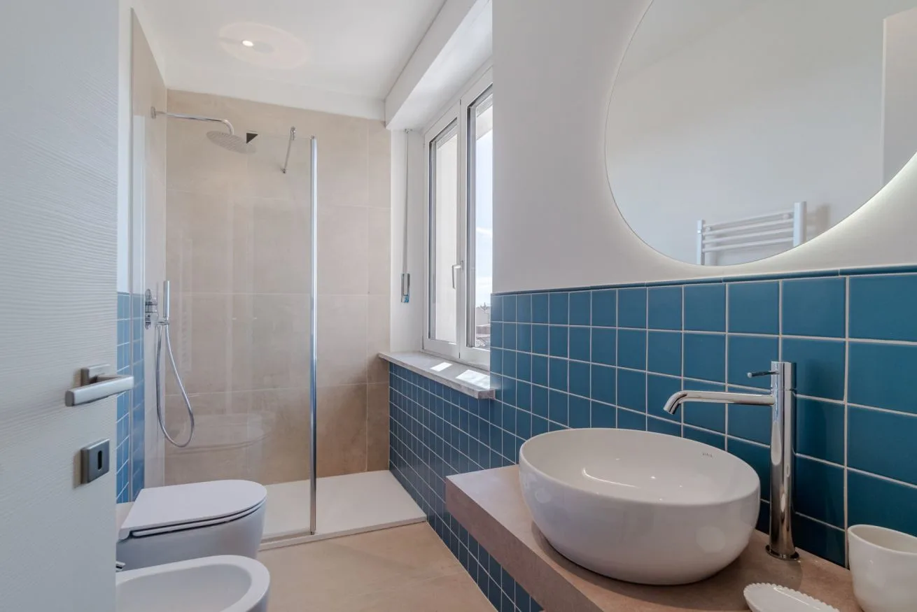 Un baño con ducha de cristal, paredes de azulejos azules y un espejo redondo grande.