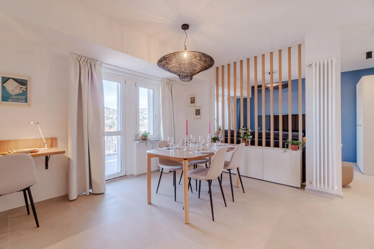 Un comedor luminoso tiene una mesa junto a ventanas altas y un separador de listones.