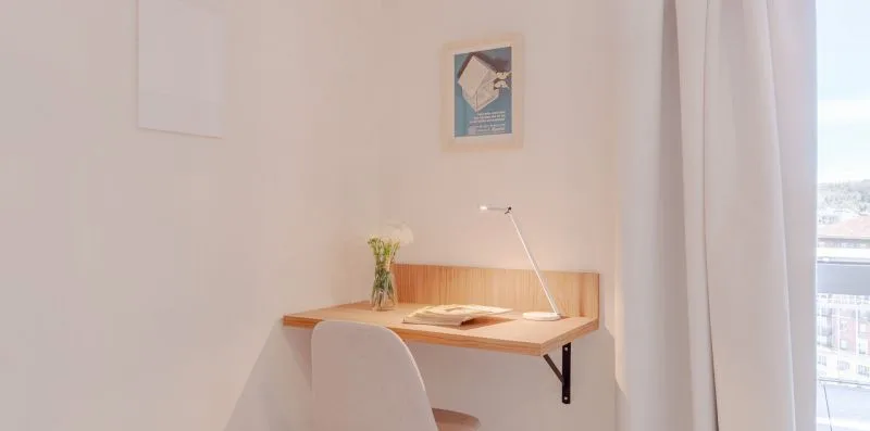 Un pequeño escritorio está junto a una pared clara, al lado de una ventana con cortinas.