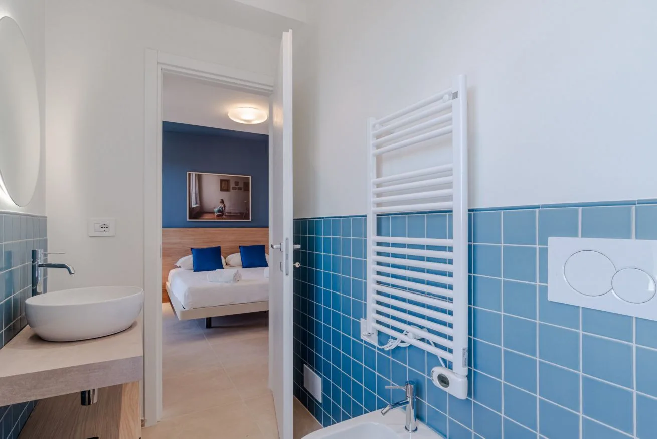 Un baño alicatado con lavabo y radiador toallero da a un dormitorio.