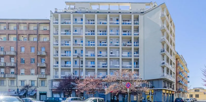 Un edificio moderno de varias plantas con filas de balcones se alza junto a una calle urbana.