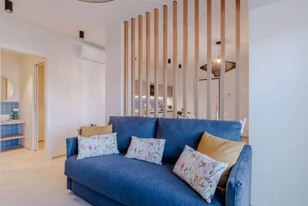 Un sofá azul con cojines estampados está junto a un separador de listones de madera en una sala luminosa.