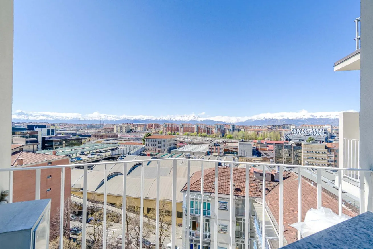 Un balcón ofrece vistas de tejados urbanos y montañas lejanas bajo un cielo azul despejado.