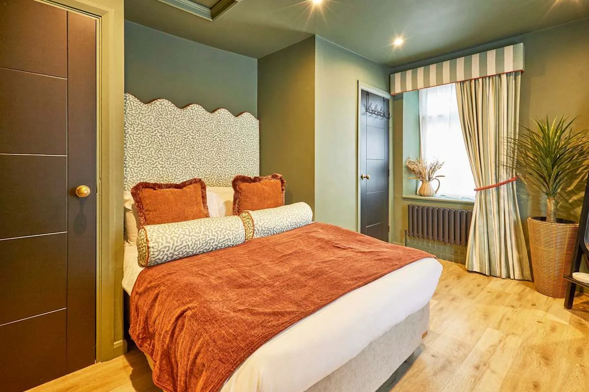 A bedroom with a bed dressed in rust-colored bedding beside a curtained window.