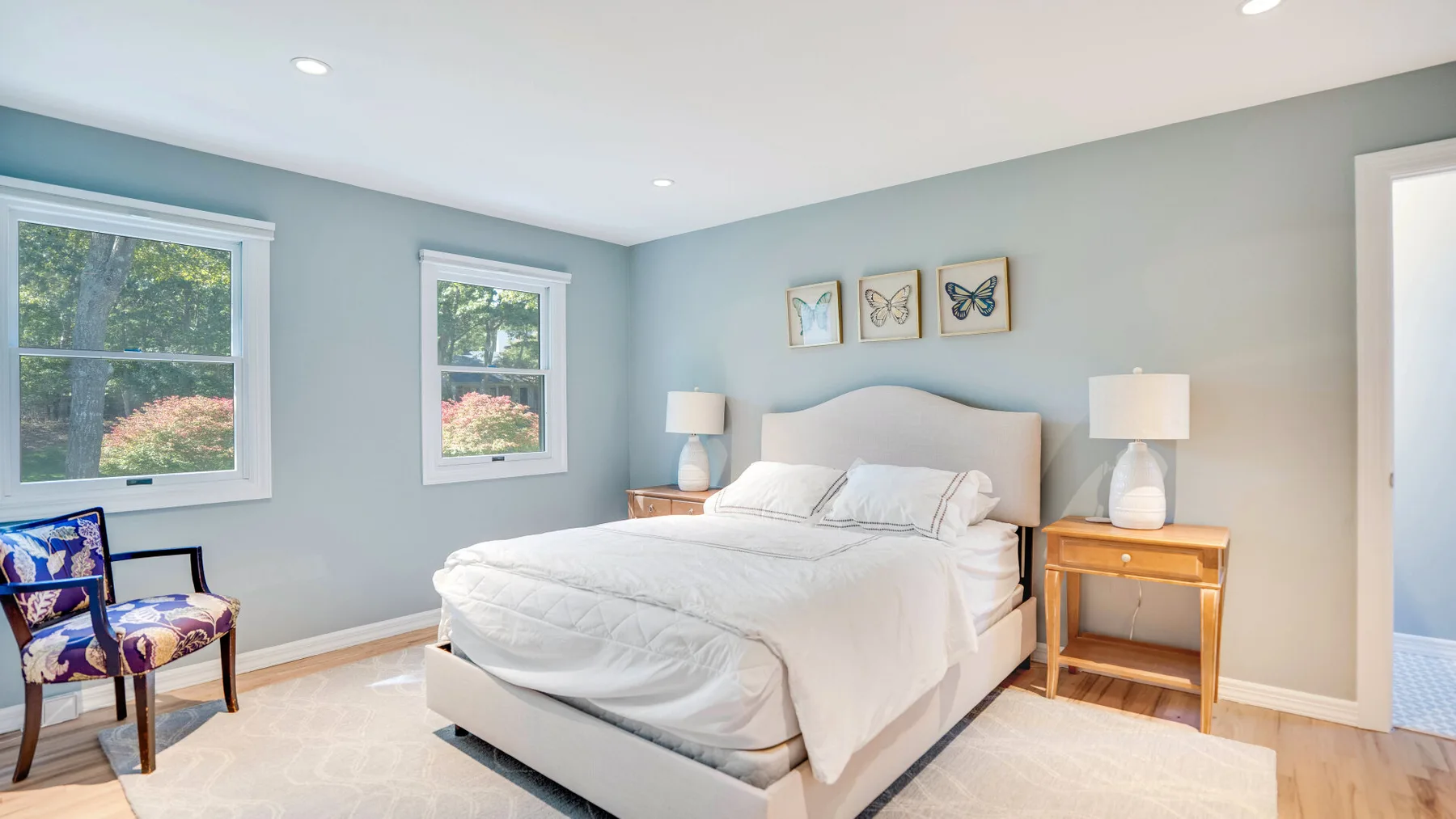 Un dormitorio luminoso con una cama bien tendida, mesitas de noche y ventanas con vistas a zonas verdes.
