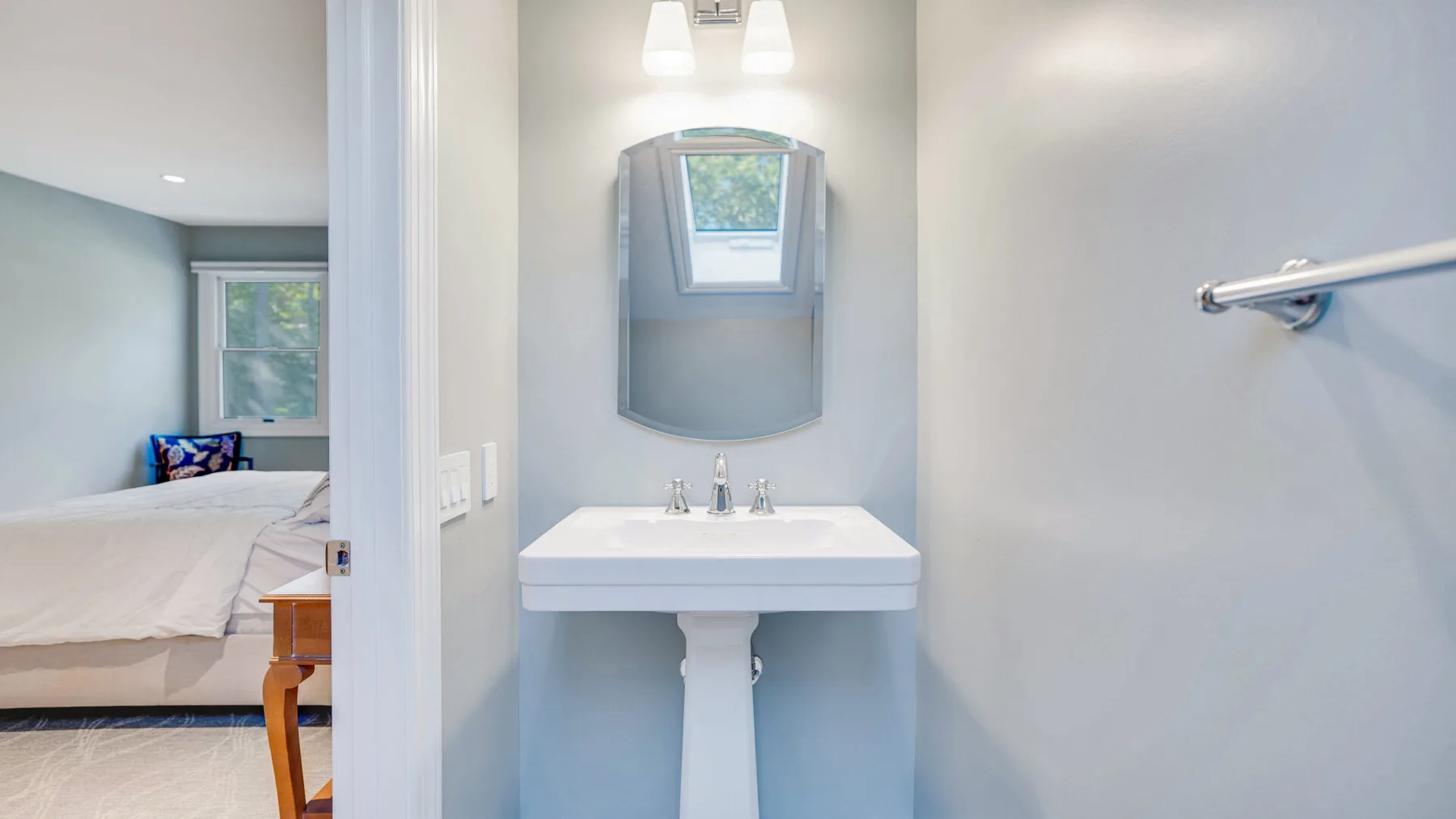 Un lavabo de baño está bajo un espejo, con un dormitorio visible por la puerta abierta.