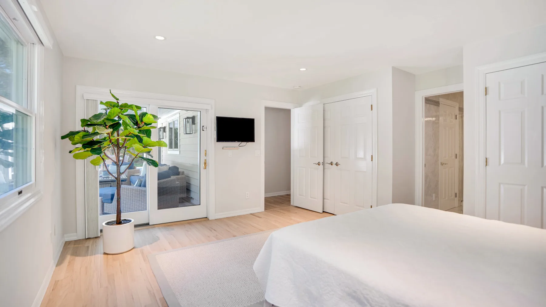 Un dormitorio luminoso con una cama blanca, suelo de madera clara y una puerta de vidrio.