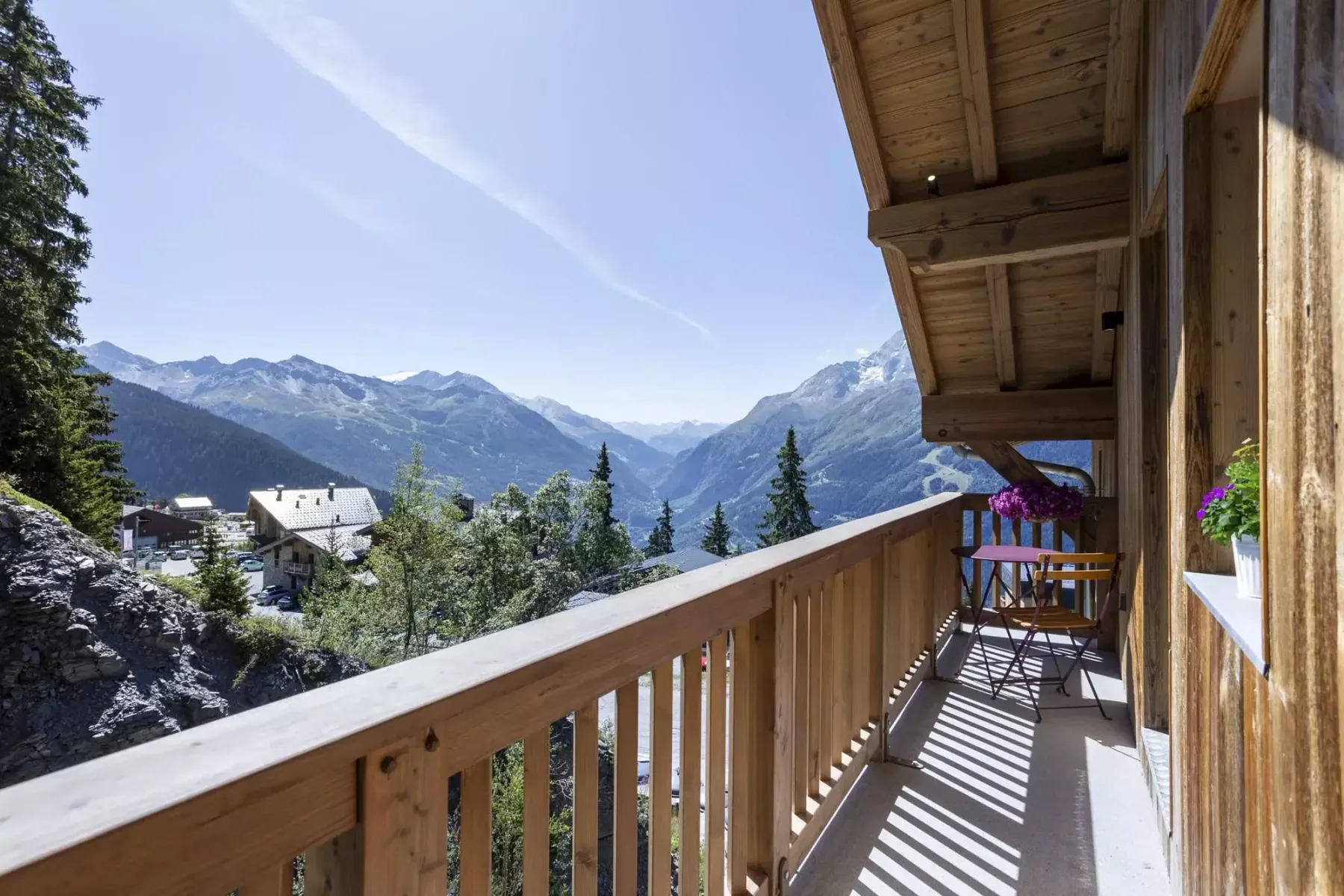 A wooden balcony overlooks a mountain valley, with outdoor seating beside the railing.