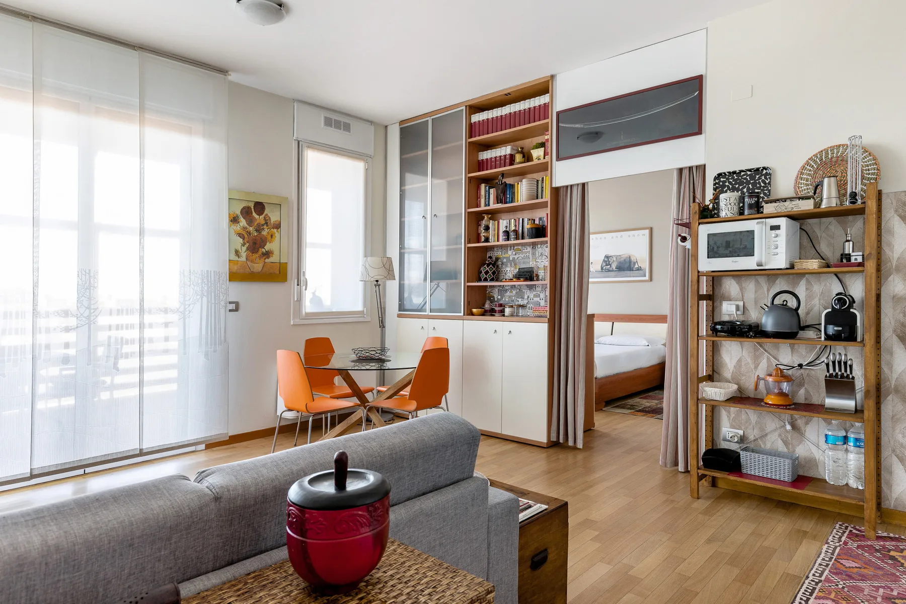 Una luminosa sala de estar tiene un sofá gris, una mesa de comedor y estanterías abiertas.