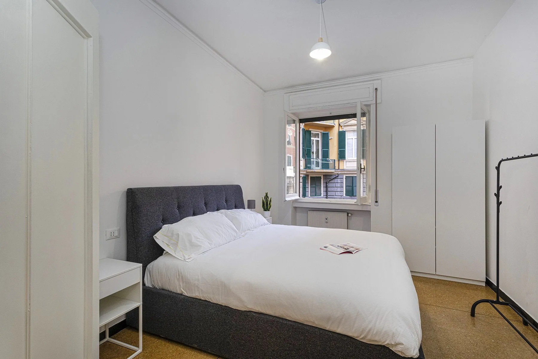 Un dormitorio luminoso con una cama bien hecha y una ventana con vista a edificios cercanos.