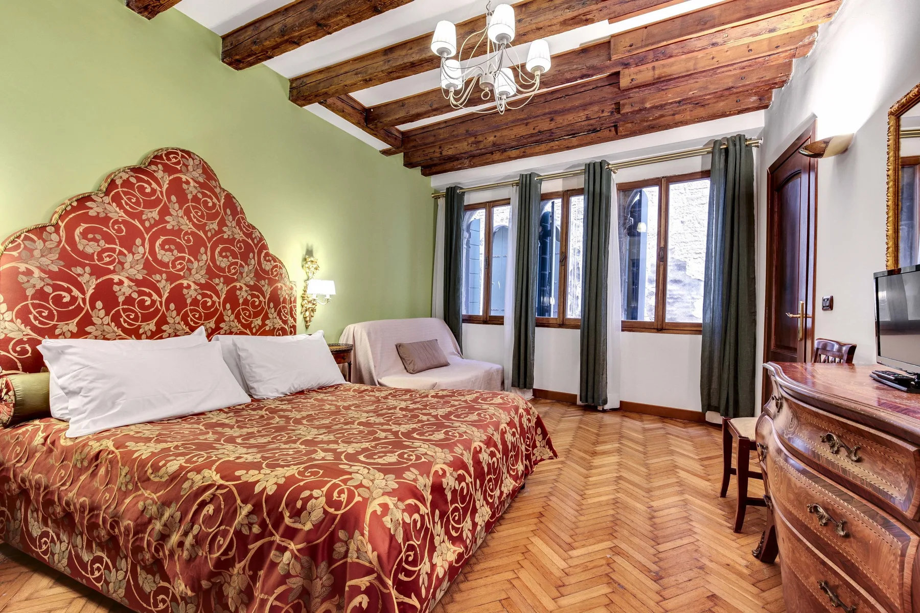 A bedroom with a patterned bedspread, a small seating area, and tall windows.