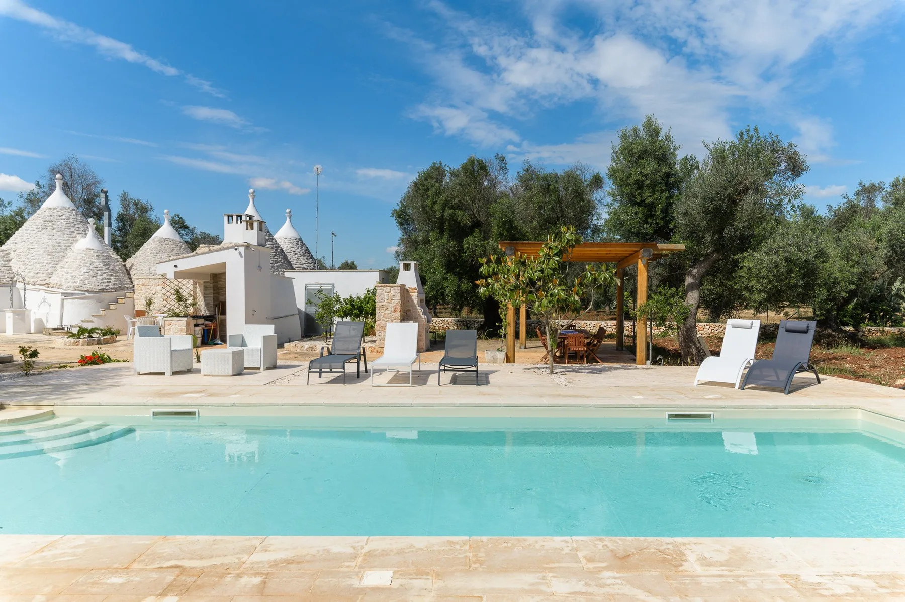 A swimming pool with lounge seating, trees, and white stone buildings in the background.
