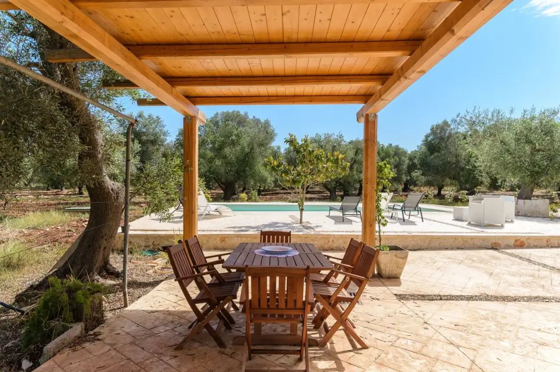 A wooden dining table and chairs sit beneath a pergola beside an outdoor pool.