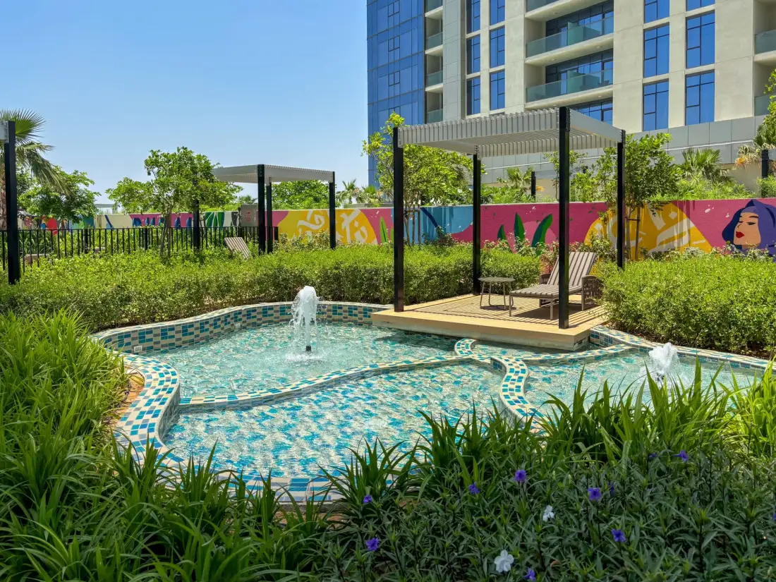 An outdoor swimming pool with fountains and a shaded seating area beside a modern building.