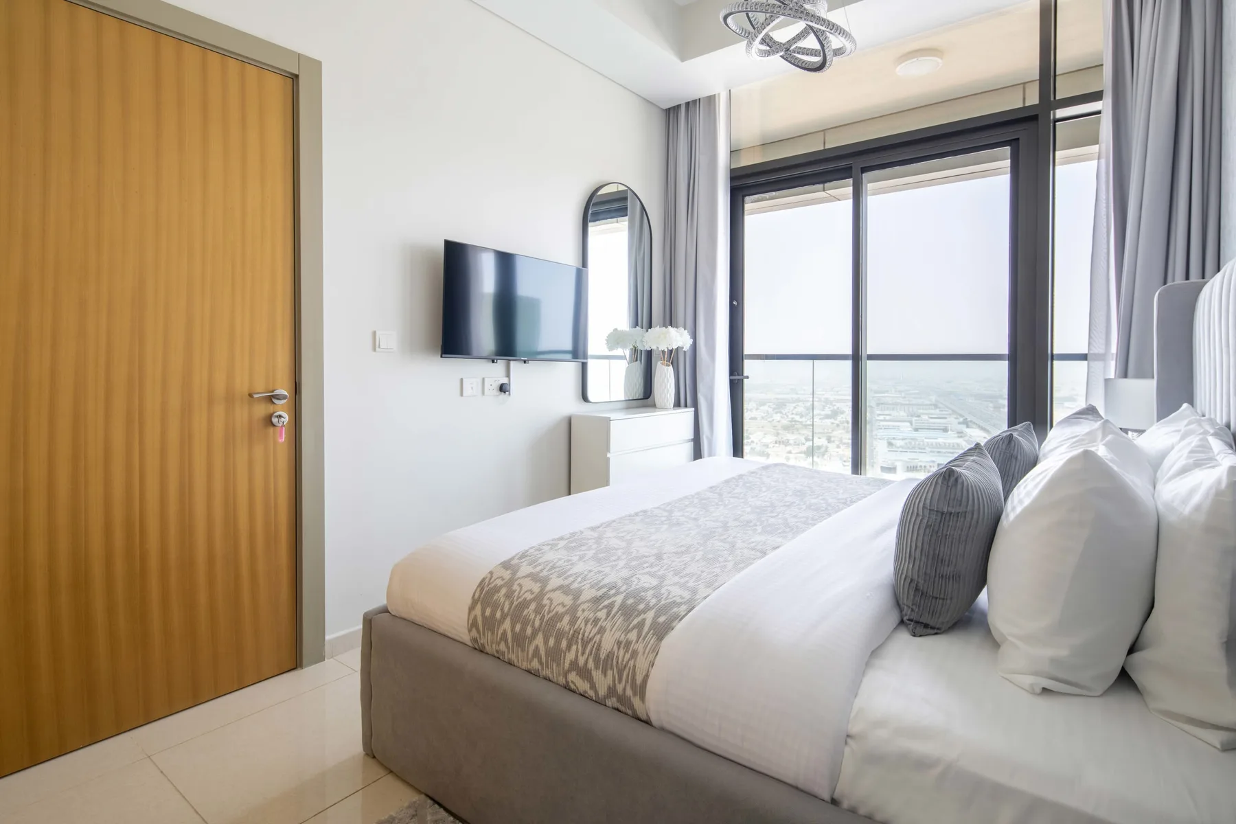 A bright bedroom with a bed beside large windows overlooking the city.