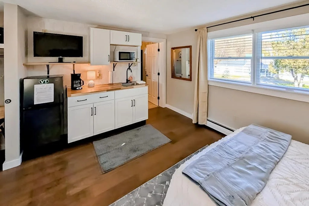 A bedroom with a bed, kitchenette, and a large window.