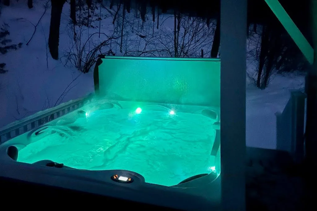 An outdoor hot tub glows turquoise beside a snowy, wooded area.