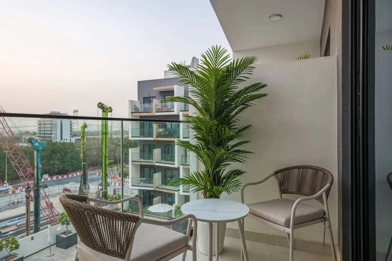 Una zona de estar en un balcón, con una mesita y una palmera en maceta, da a edificios cercanos.