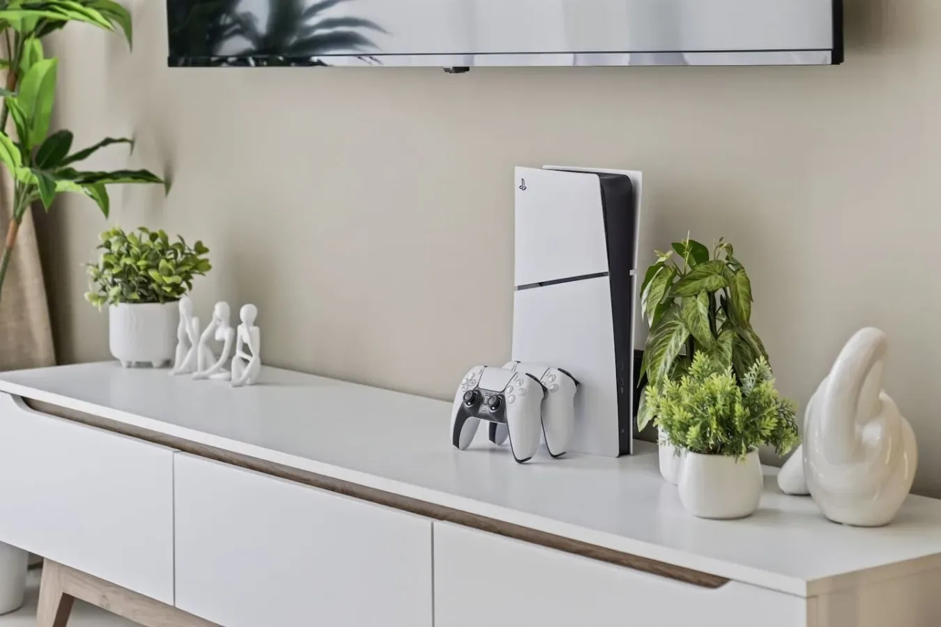 Un mueble blanco para medios sostiene una consola de videojuegos, un control, plantas y figuras decorativas.