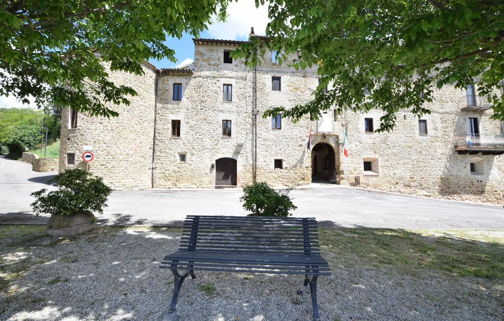 A stone building stands behind a bench beneath leafy trees.