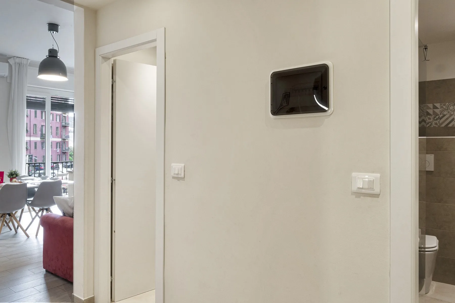 A hallway wall with a small recessed screen, with a dining area and bathroom visible nearby.