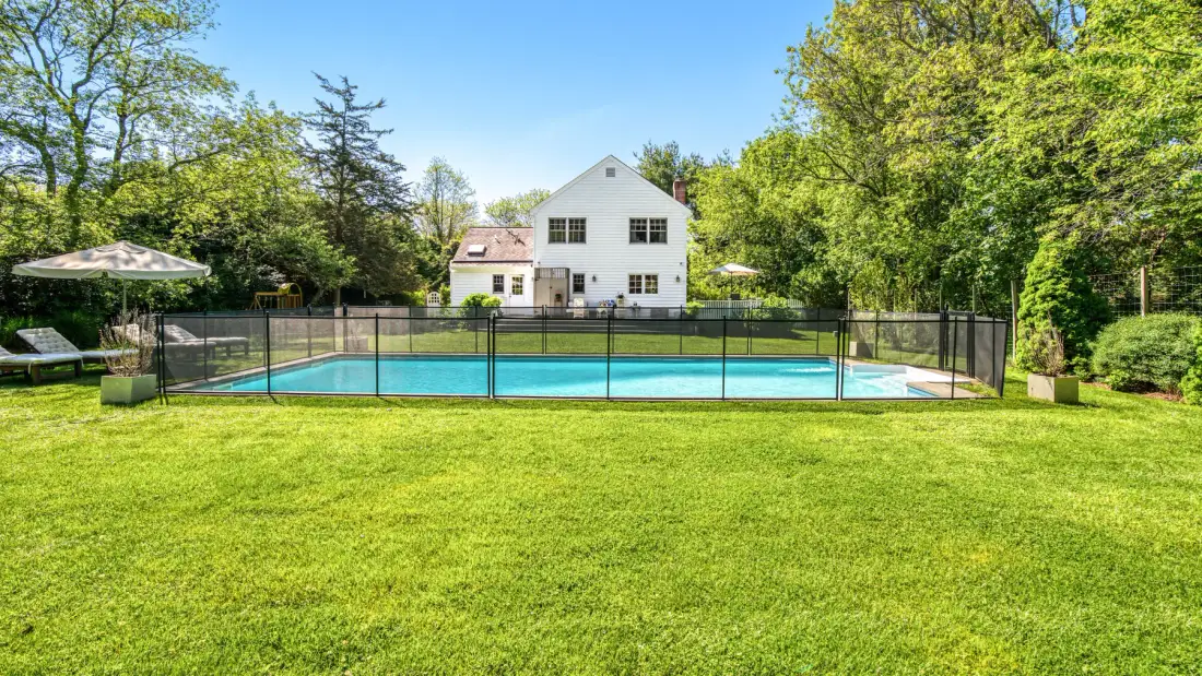 Una piscina exterior cercada está sobre un amplio césped verde frente a una casa.