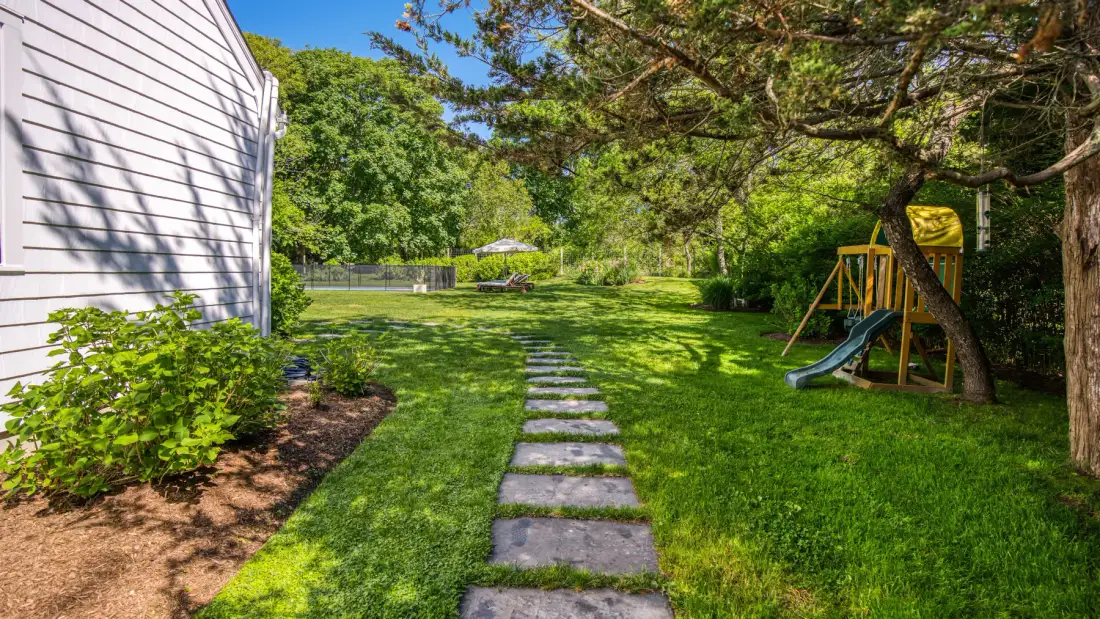Un patio con césped, un sendero de piedras, árboles y una pequeña estructura de juego.