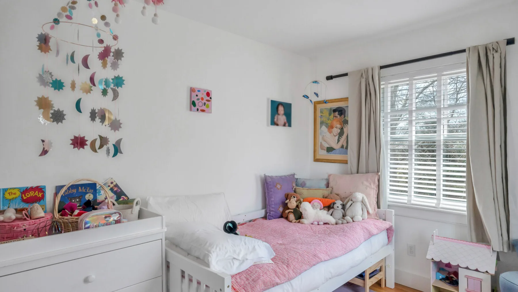 Un dormitorio luminoso con una cama cubierta de rosa, juguetes y una gran ventana con persianas.