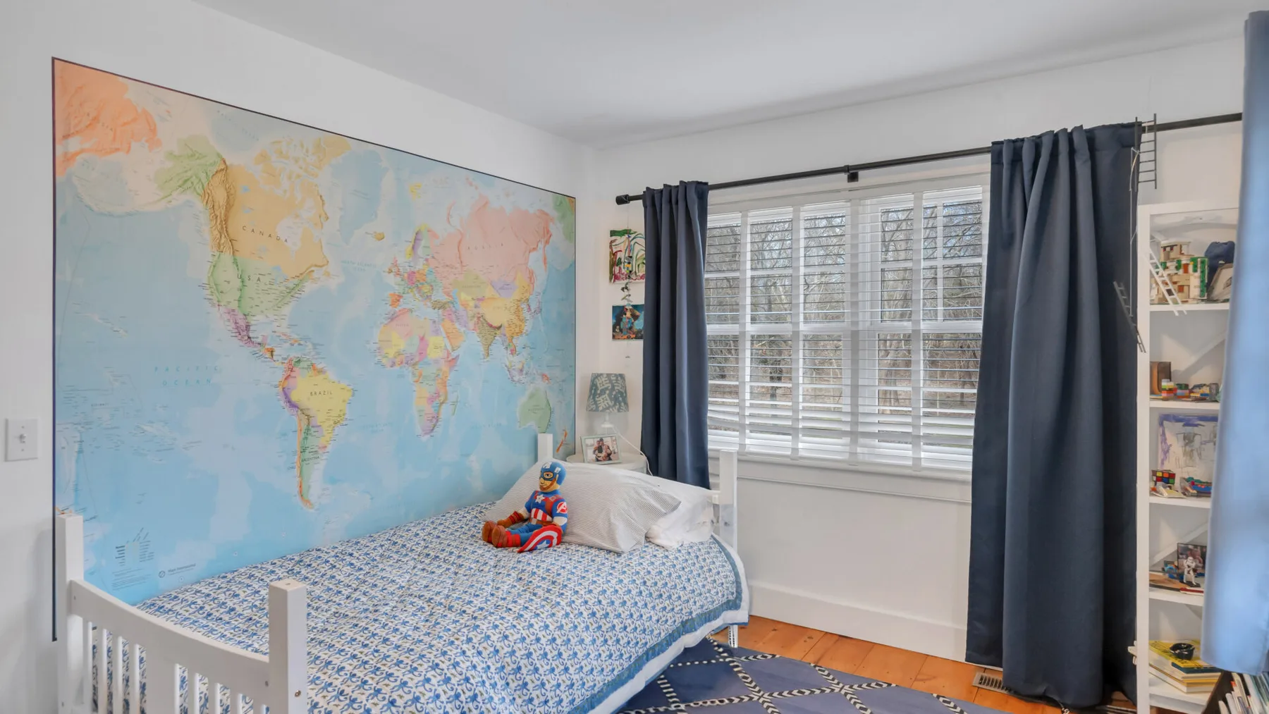 Un dormitorio con una cama bajo un mapa mundial colorido y una ventana con cortinas oscuras.