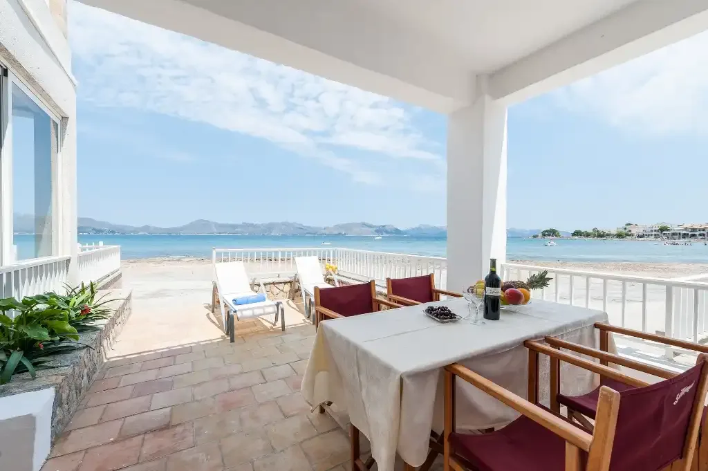 A seaside patio with a dining table and chairs overlooks the beach and water.