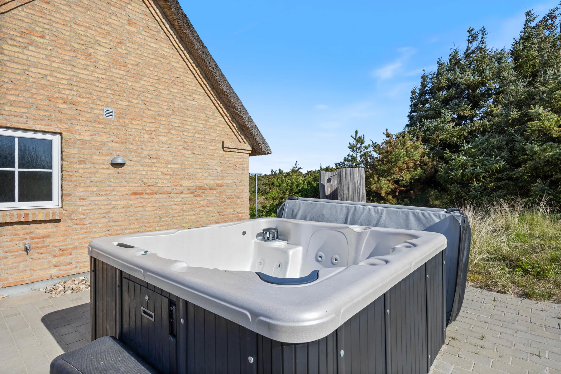 Una bañera de hidromasaje exterior está junto a una casa de ladrillo, con árboles y césped al fondo.