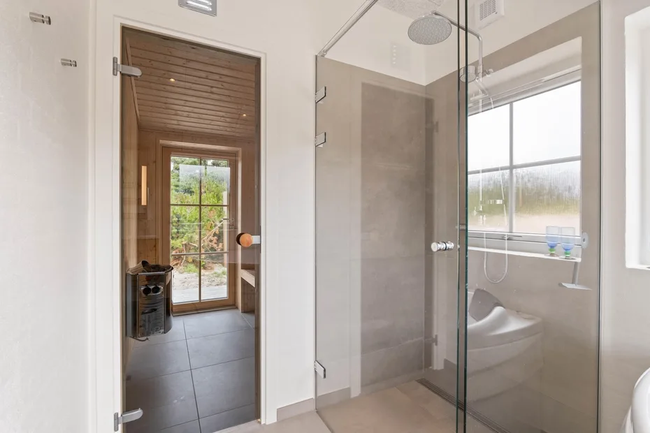 Una ducha de baño con mampara de vidrio está junto a una ventana con vista a la vegetación.