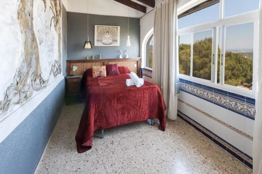 A bedroom with a red bed beside large windows overlooking trees.