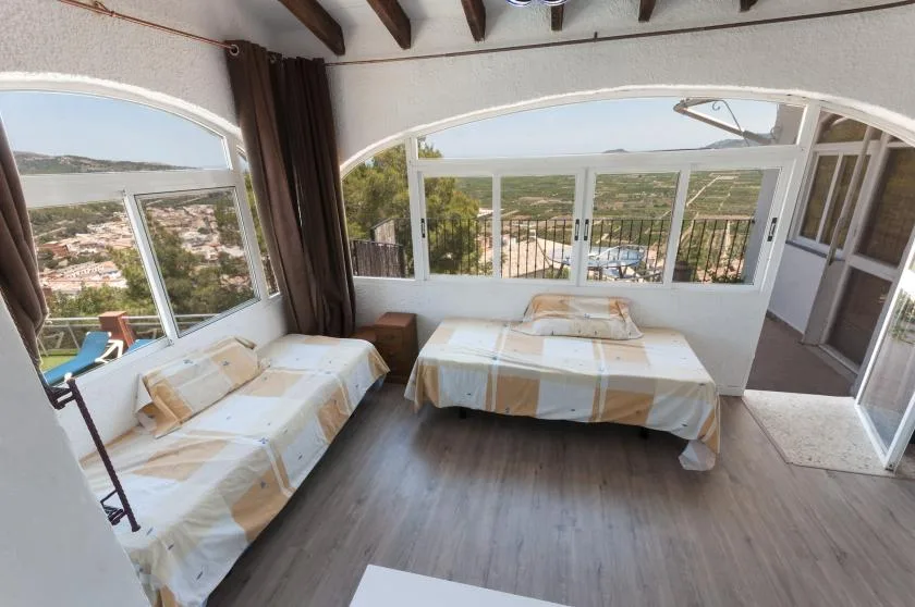 A bright bedroom with two beds beside large windows overlooking a hillside.