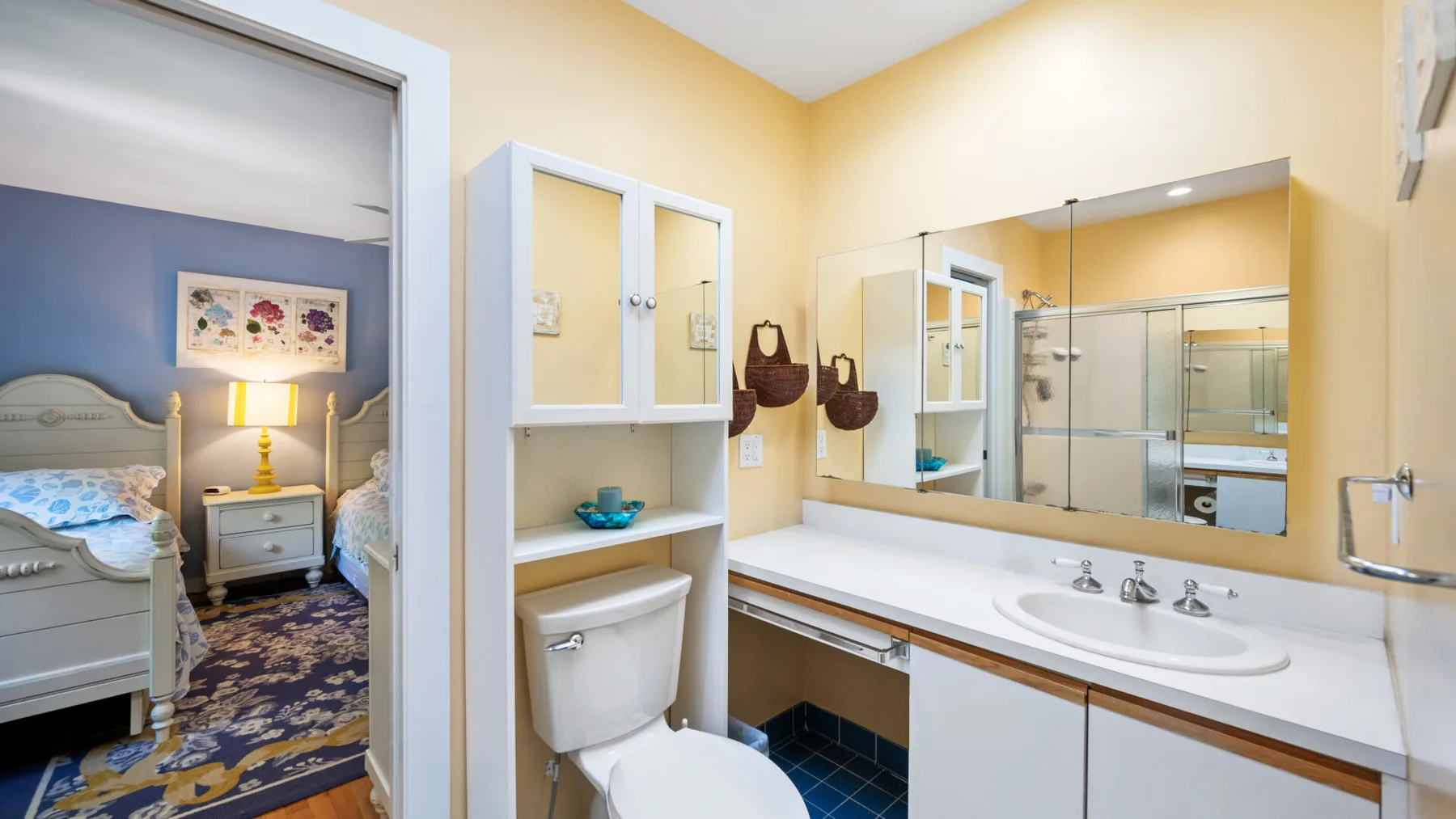 A bathroom with a sink, toilet, and large wall mirror beside a bedroom.