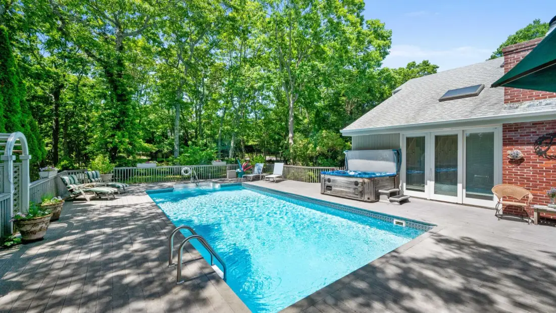 A backyard swimming pool beside a house, surrounded by trees and patio seating.