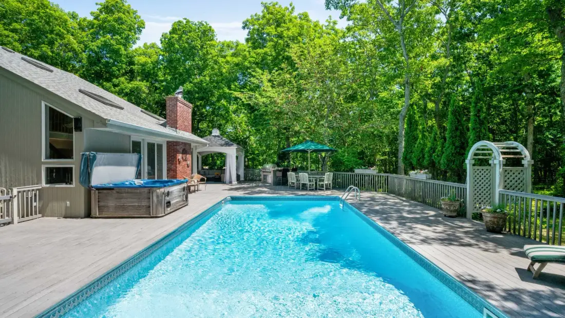 A swimming pool and surrounding deck sit beside a house, with trees beyond.