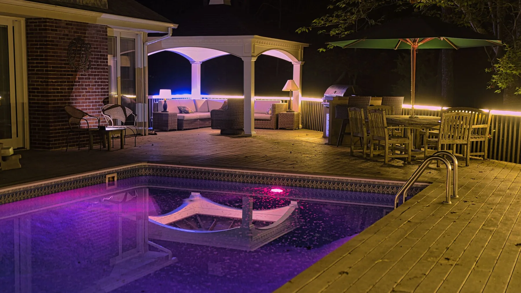 An outdoor pool and wooden deck are illuminated at night beside a covered seating area.