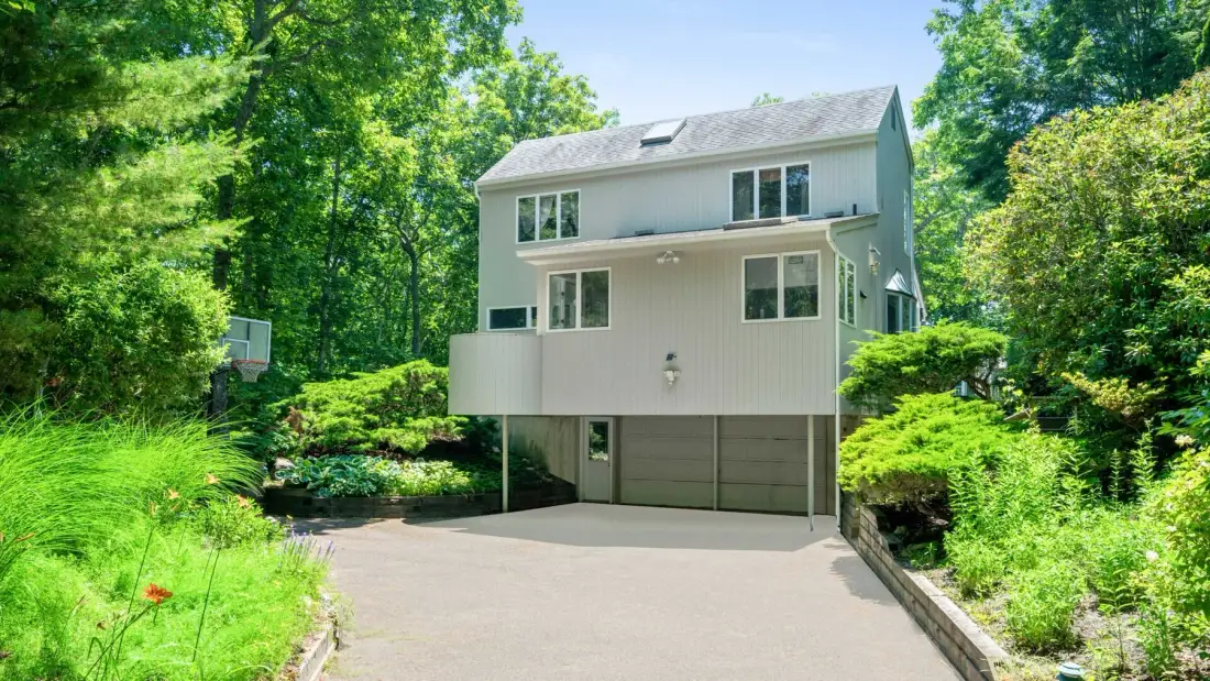 A two-story house with a wide driveway, surrounded by trees and greenery.