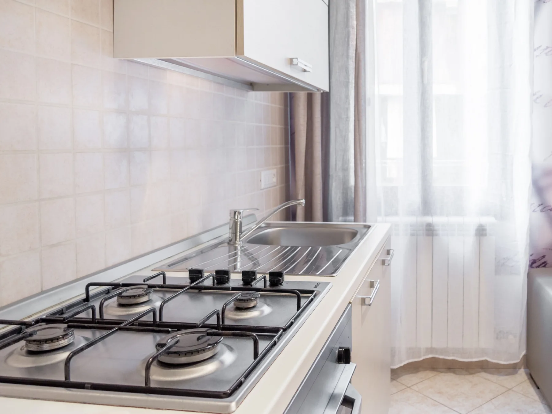 A kitchen with a gas cooktop, sink, and a window with sheer curtains.
