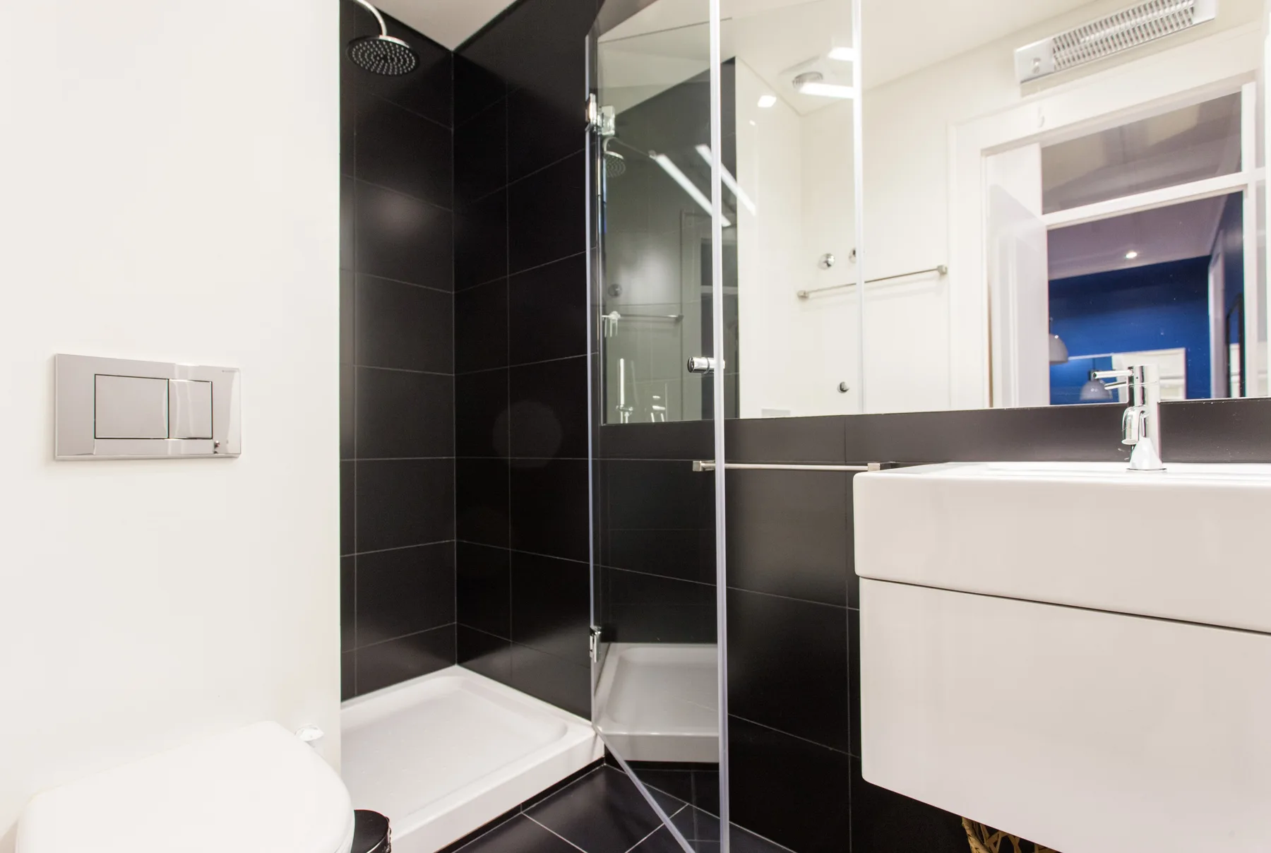 Un baño con ducha de vidrio, lavabo suspendido y azulejos negros.