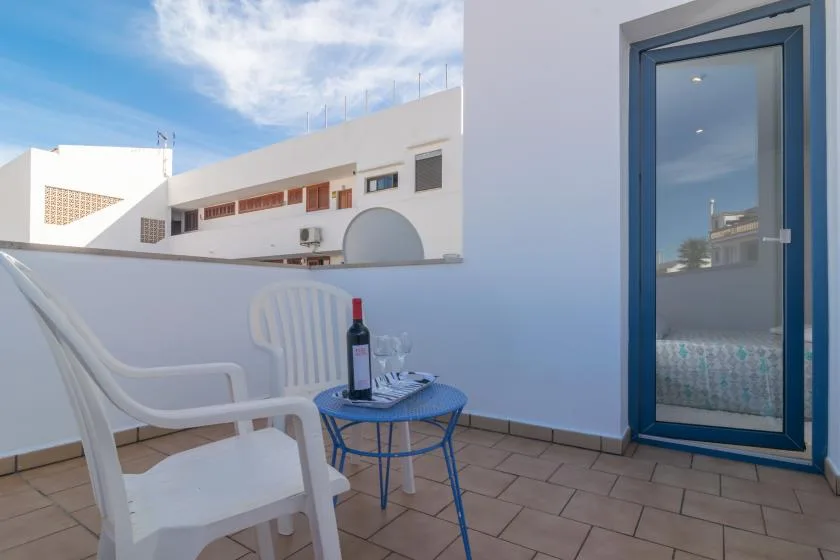 A small outdoor terrace has white seating, a blue table, and a glass door.
