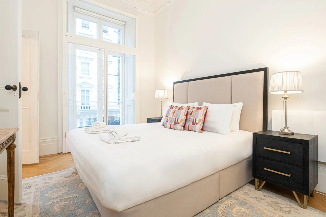 A bright bedroom with a large bed, bedside tables, and tall windows.