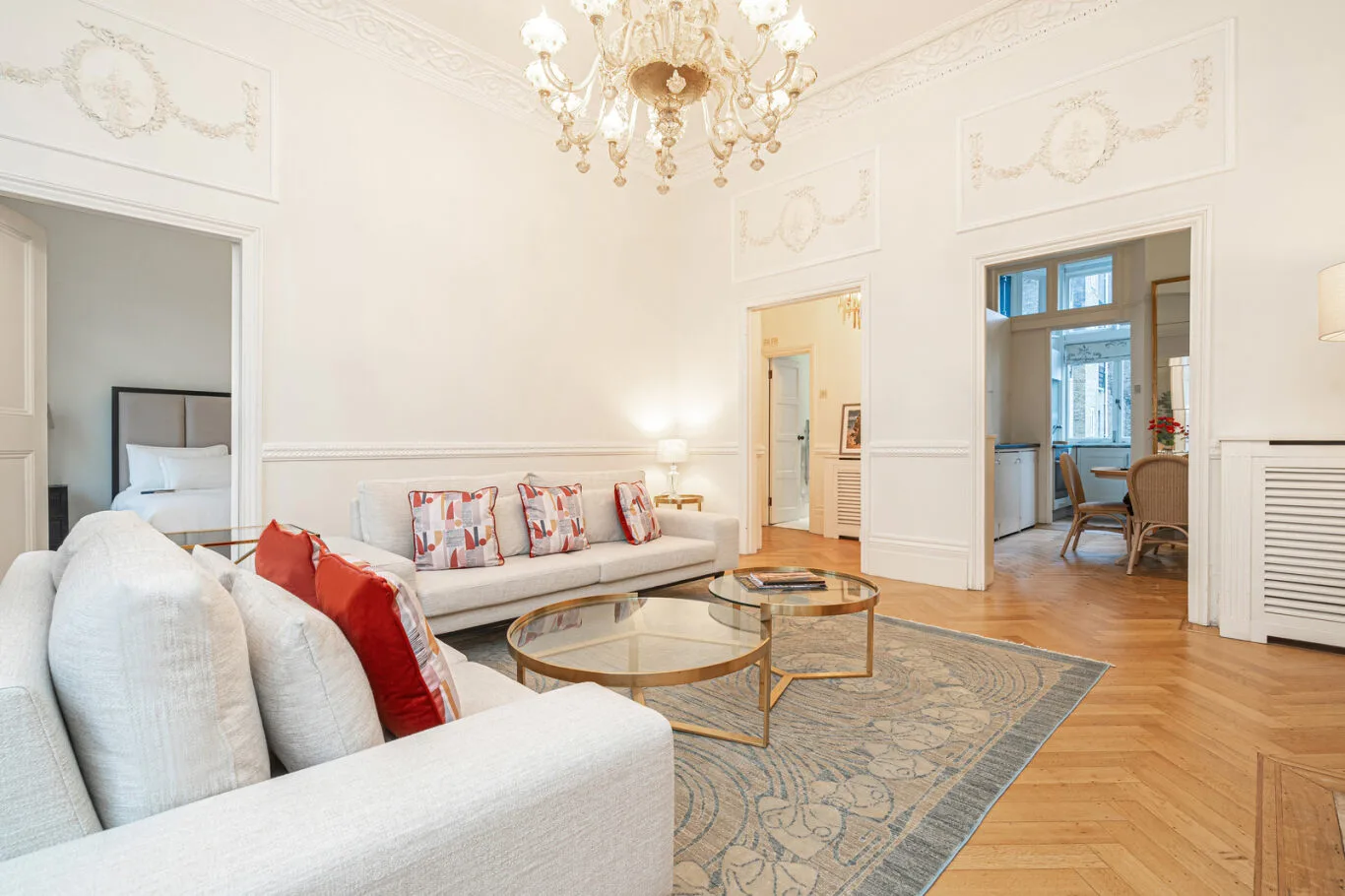 A bright living room with pale seating, glass coffee tables, and a patterned rug.