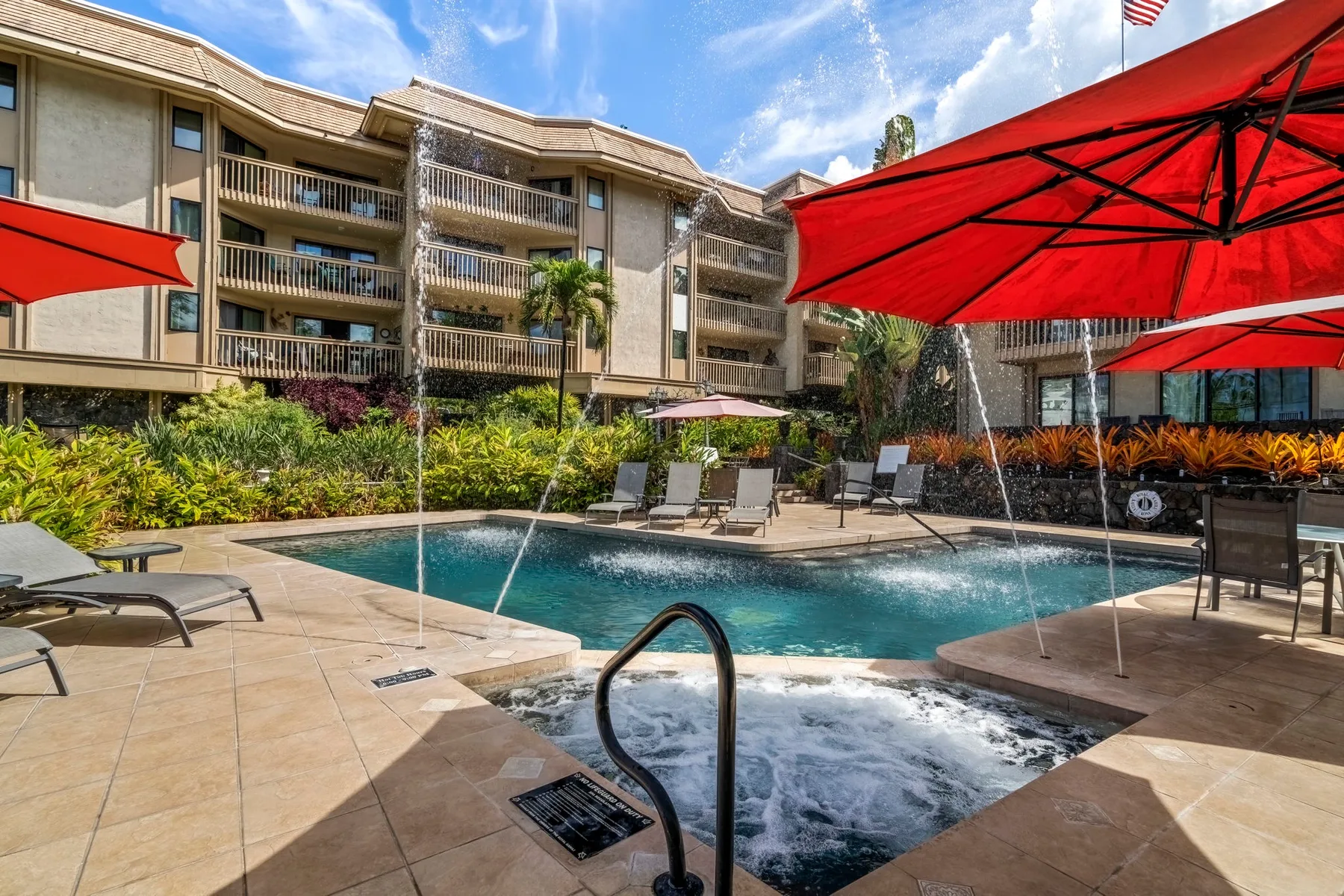 An outdoor pool and adjacent spa are surrounded by lounge seating and red umbrellas.