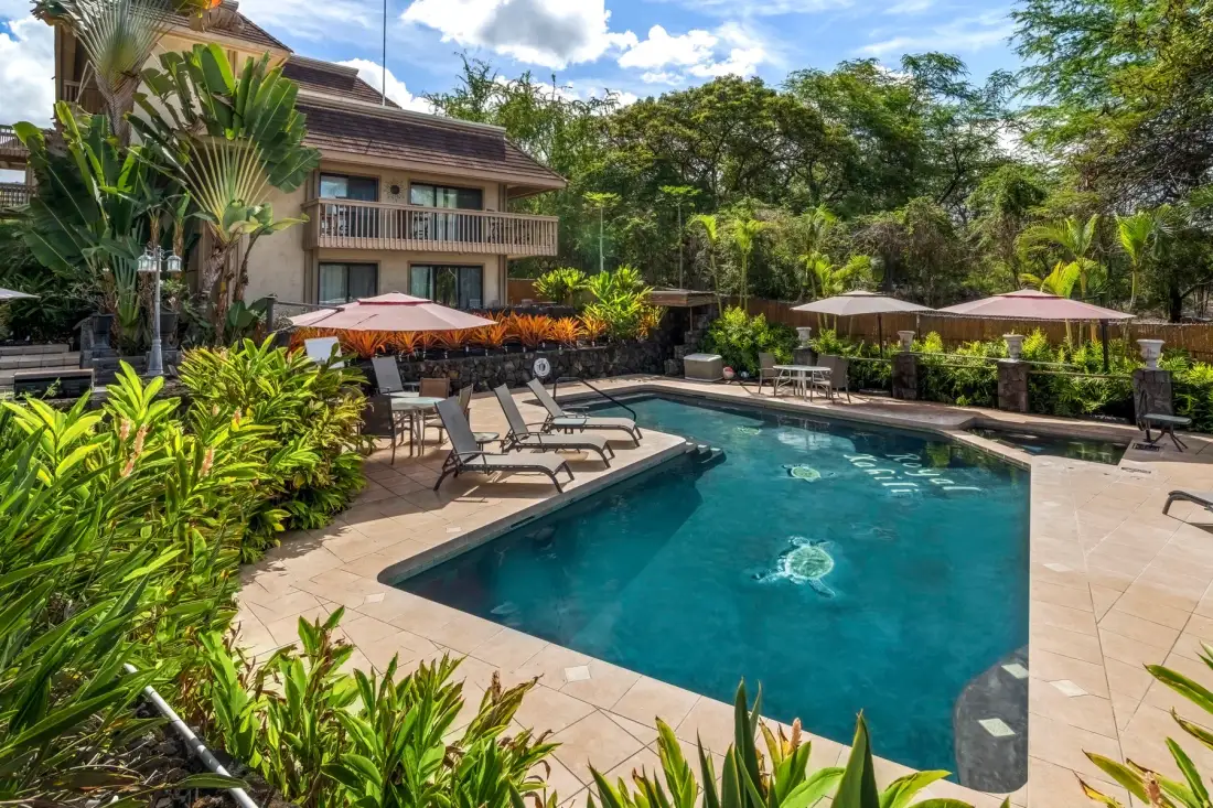 An outdoor swimming pool is surrounded by lounge seating and tropical plants.