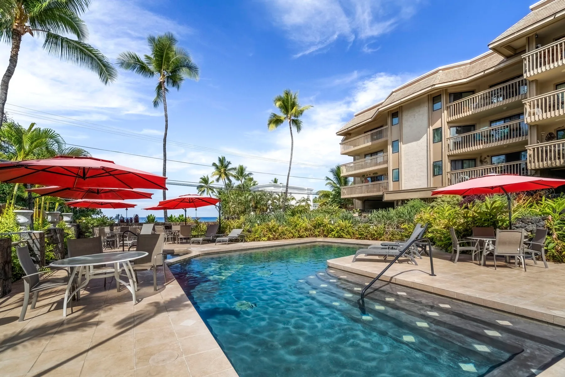 An outdoor swimming pool beside a multi-story building, with patio seating and red umbrellas.