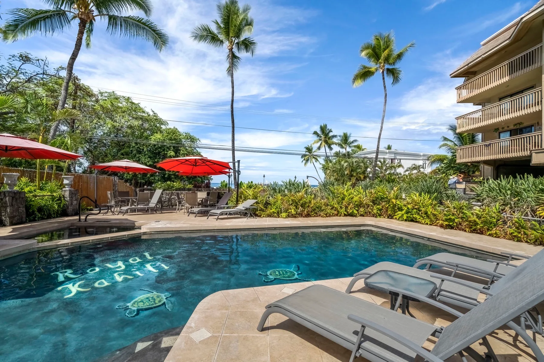 An outdoor swimming pool is surrounded by lounge chairs, umbrellas, and palm trees.