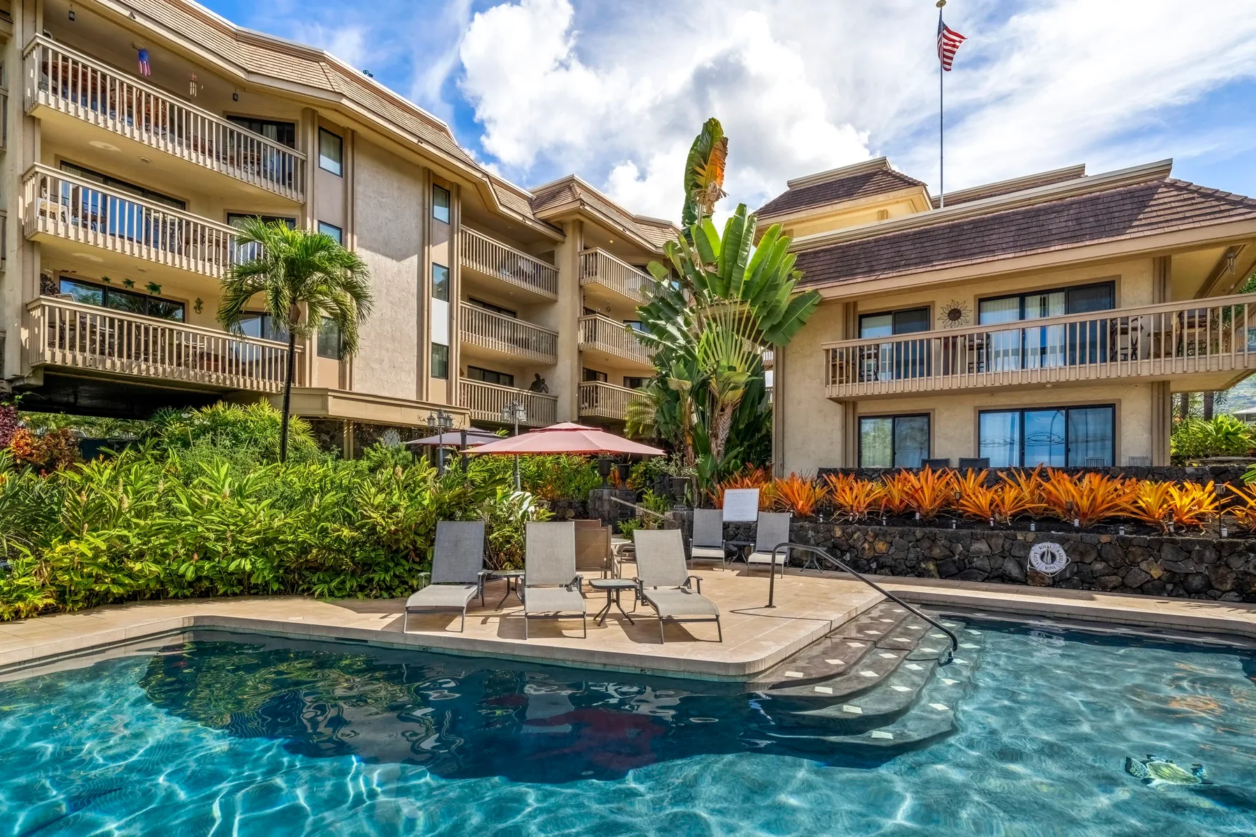 An outdoor swimming pool sits beside a multi-story building and landscaped garden.