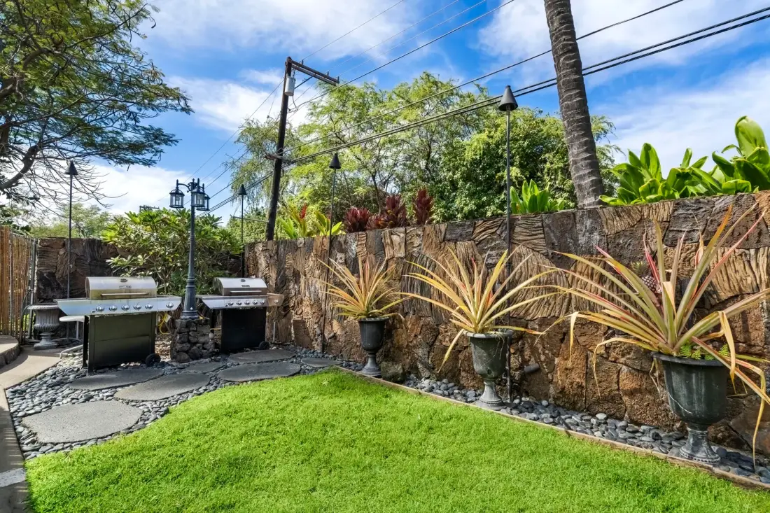 An outdoor garden area with a lawn, potted plants, and grills beside a stone wall.